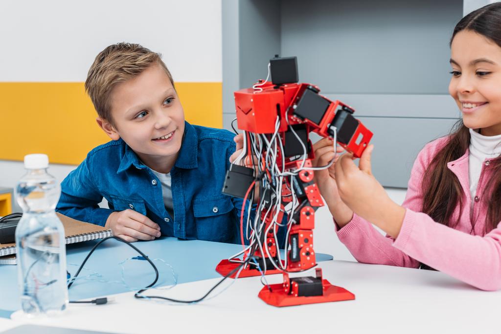 ihana koulutyttö ja hymyilevä koulupoika istuu pöydän ääressä ja työskentelee yhdessä robotin mallin kanssa STEM-luokassa
 - Valokuva, kuva