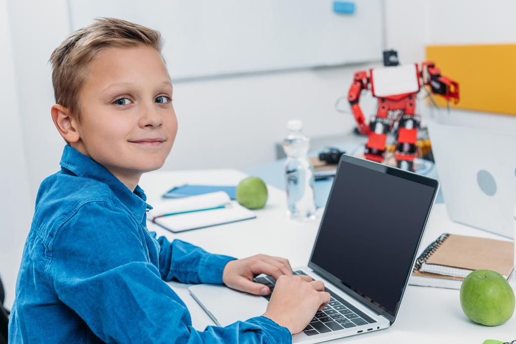 schoolboy sitting at table with robot model, looking at camera and using laptop with blank screen during STEM lesson   - Photo, Image