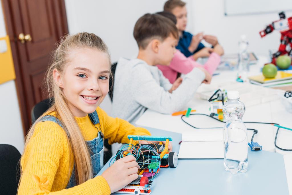 adorable schoolgirl holding robot model, looking at camera while classmates having STEM lesson - Photo, Image