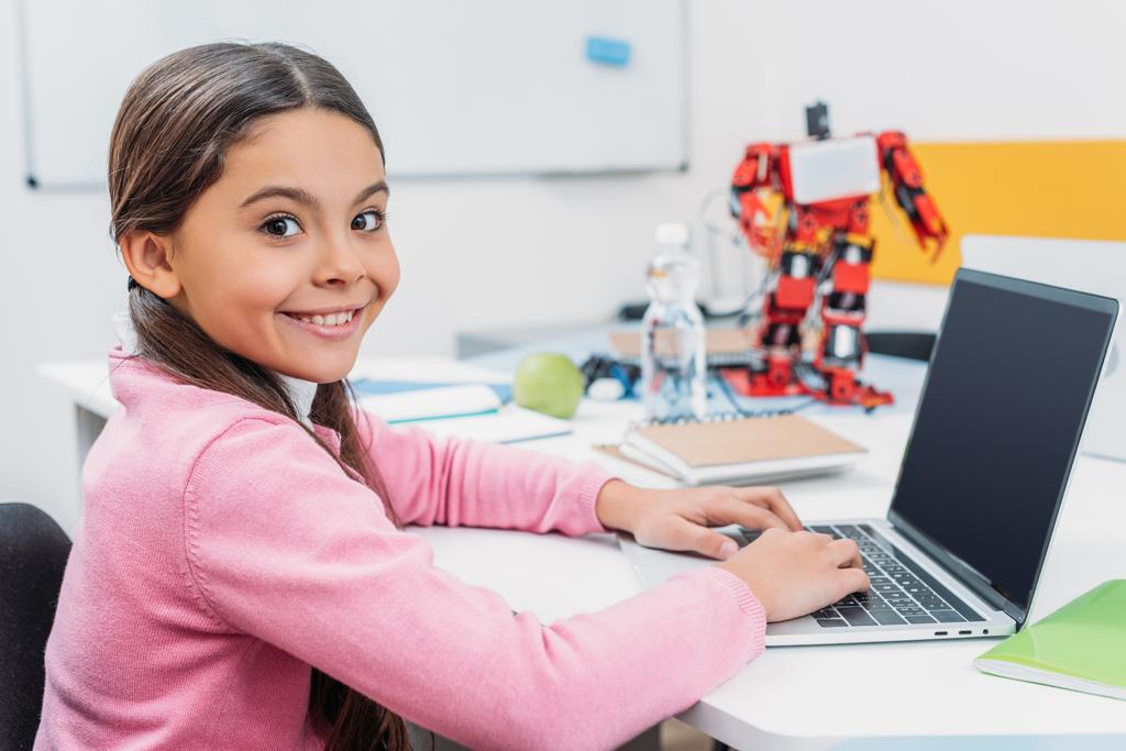 adorable schoolgirl sitting at table with robot model, looking at camera and using laptop with blank screen during STEM lesson   - Photo, Image