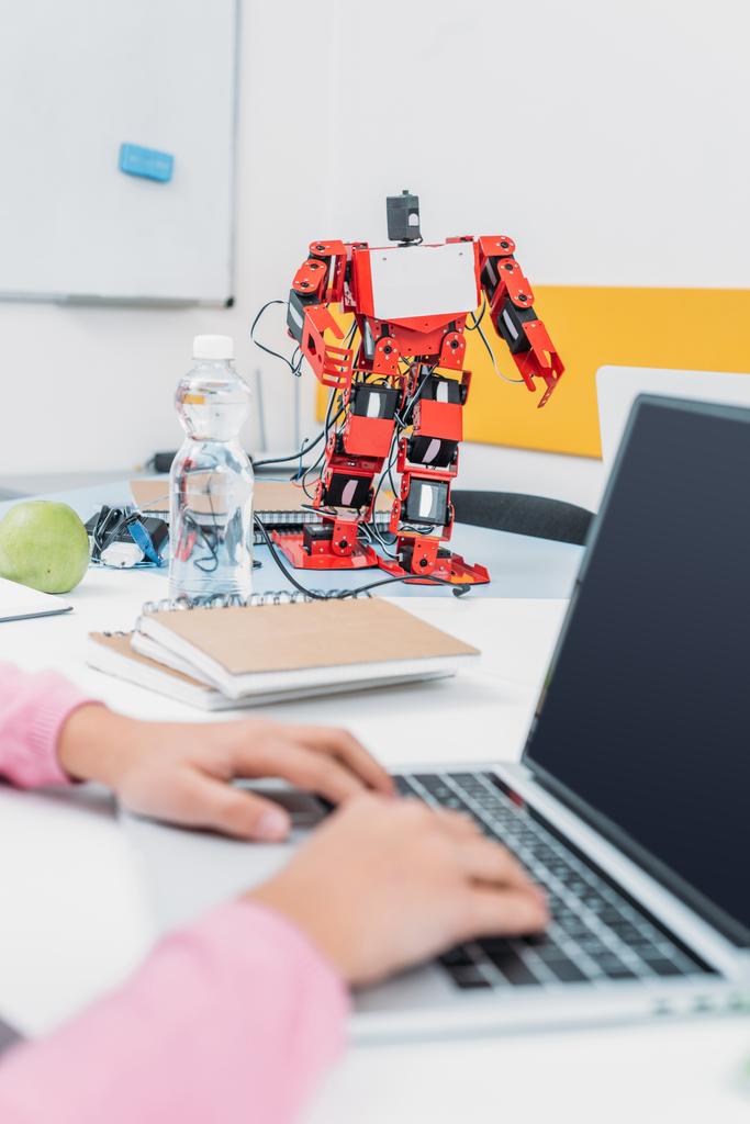 oříznutý pohled školačka sedí u stolu s modelem robota a pomocí přenosného počítače s prázdnou obrazovkou během lekce stonku   - Fotografie, Obrázek