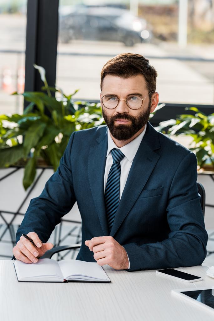 confident buisnessman in eyeglasses sittting at workplace and looking at camera - Photo, Image