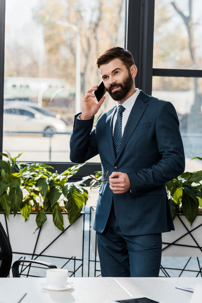 Улыбающийся бородатый бизнесмен в формальной одежде разговаривает со смартфоном в офисе
 - Фото, изображение