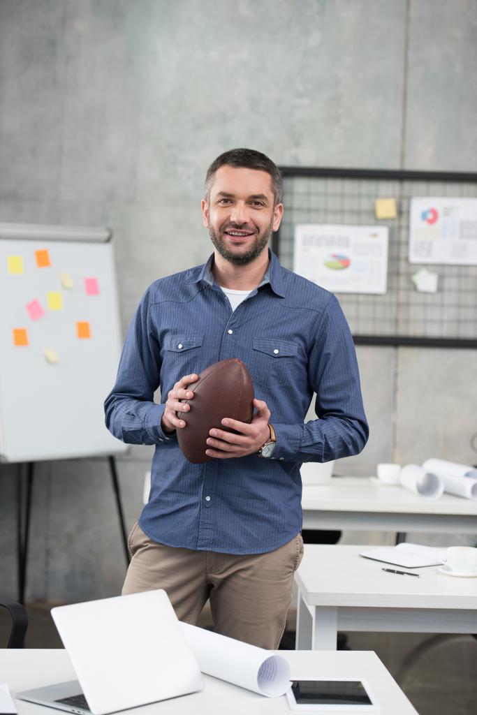 uśmiechający się przystojny biznesmen trzymając piłkę futbolu amerykańskiego i patrząc na kamery w biurze - Zdjęcie, obraz