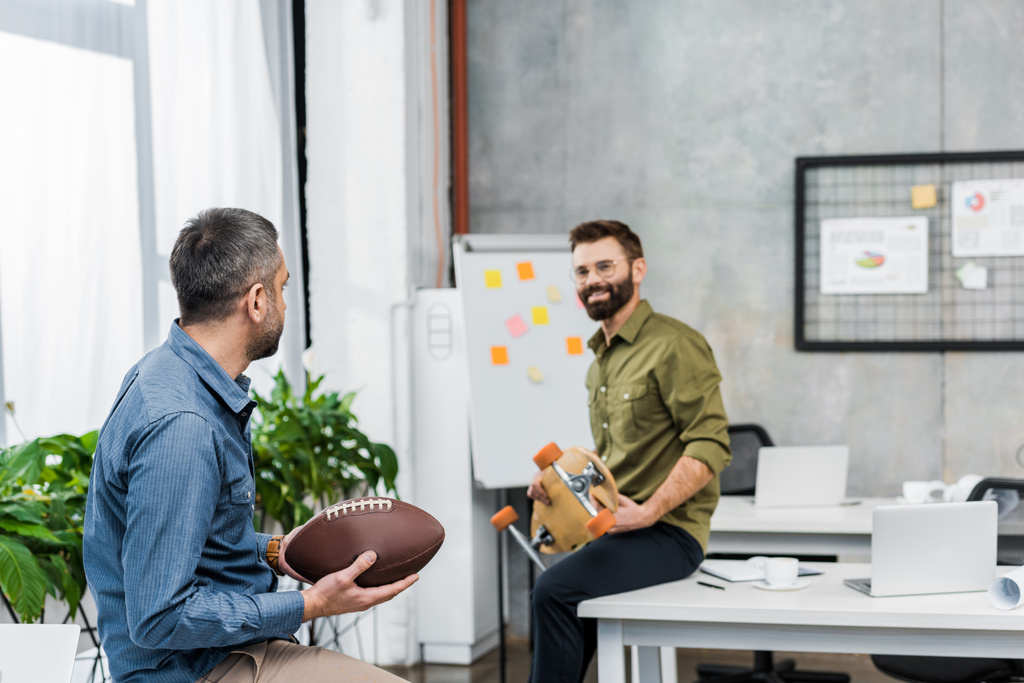 biznesmeni uśmiechający się patrząc na siebie nawzajem i trzymając piłkę futbolu amerykańskiego i longboard w biurze - Zdjęcie, obraz