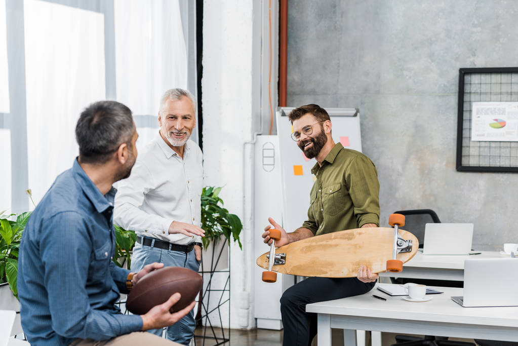 Красивые бизнесмены разговаривают и держат в офисе скейтборд и американский футбольный мяч
 - Фото, изображение