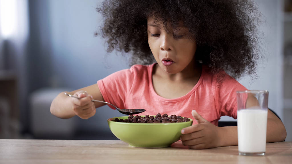 Cute mulatto girl eating chocolate cornflakes for breakfast, risk of diabetics - Photo, Image