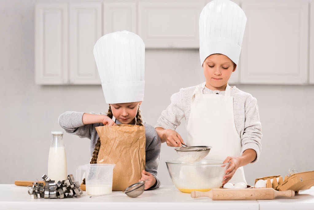 entzückende Kinder in Schürzen und Kochmützen bei der Essenszubereitung am Tisch in der Küche  - Foto, Bild