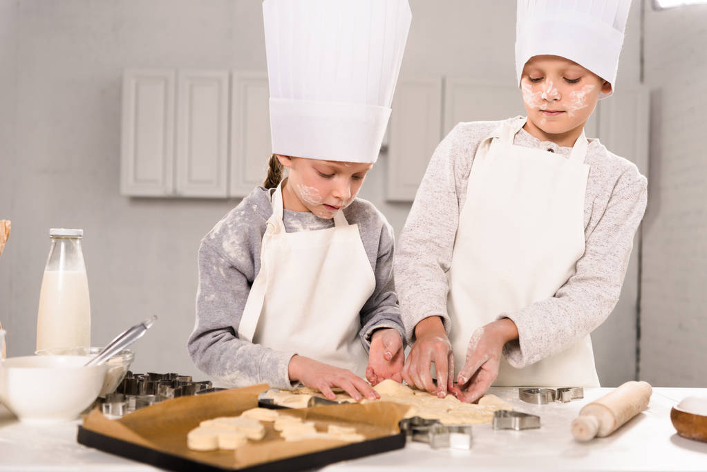 niños en gorros de chef y delantales cortando masa para galletas en la mesa en la cocina
 - Foto, Imagen