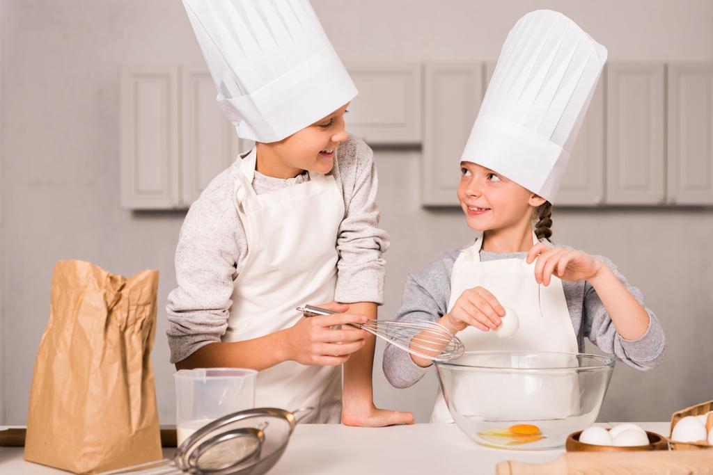 irmã sorridente e irmão em chapéus de chef e aventais batendo ovos na tigela na mesa na cozinha
 - Foto, Imagem