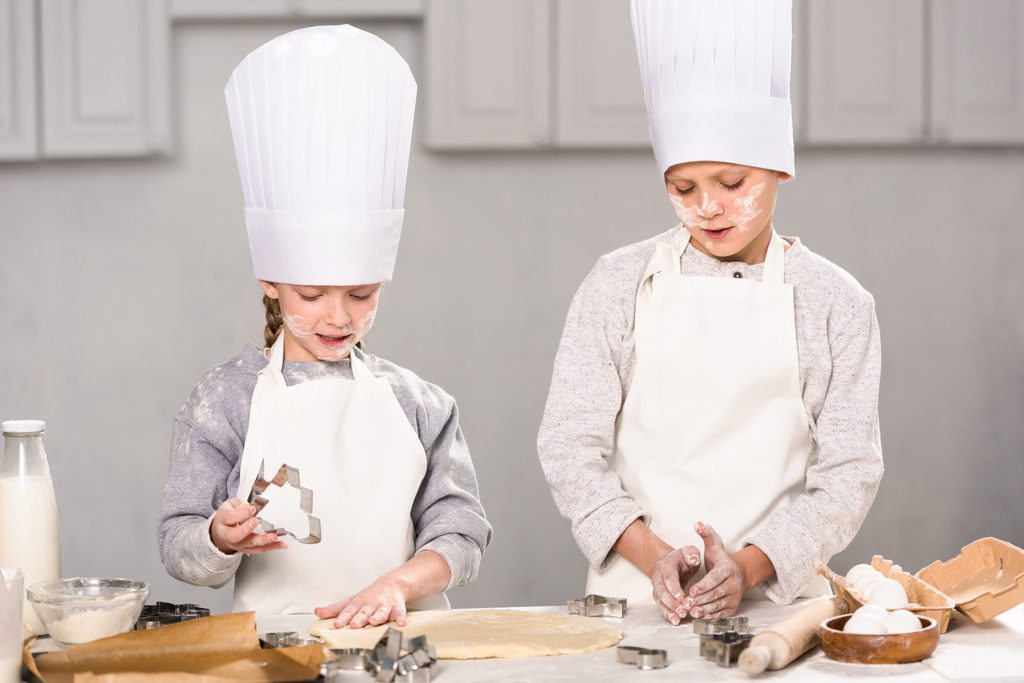 Kinder in Kochmützen und Schürzen schneiden Teig für Plätzchen am Tisch in der Küche aus - Foto, Bild