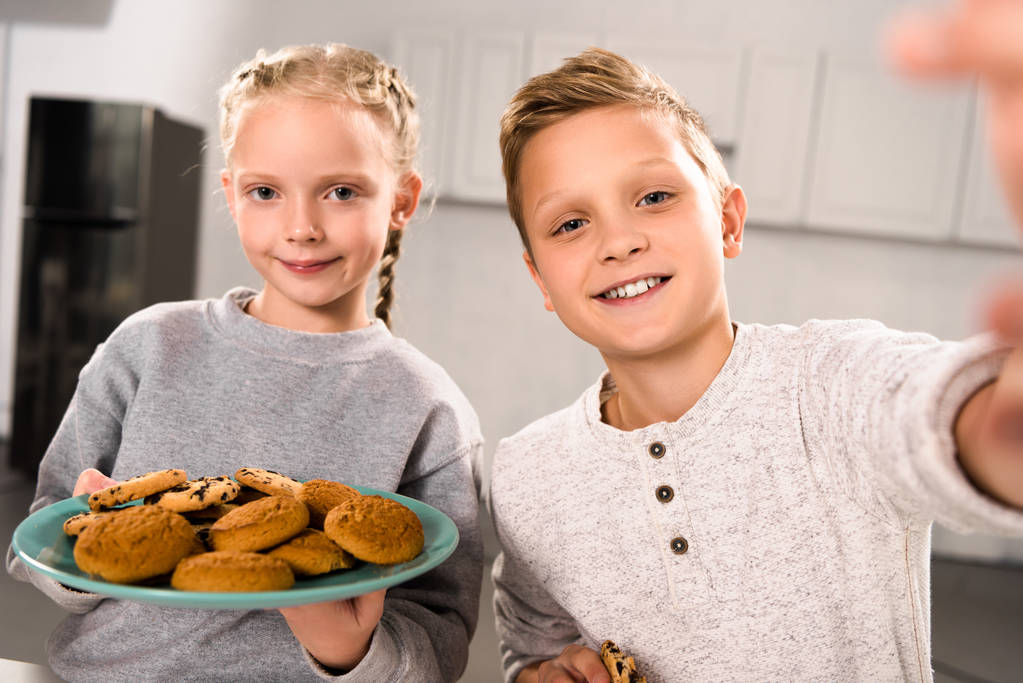 foco seletivo do menino tomando selfie com a irmã enquanto ela segurando prato com biscoitos
 - Foto, Imagem