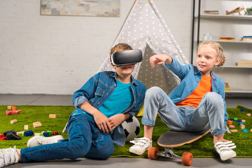 niño usando auriculares de realidad virtual y su hermana señalando con el dedo mientras está sentado en el monopatín cerca de wigwam en casa
 - Foto, imagen
