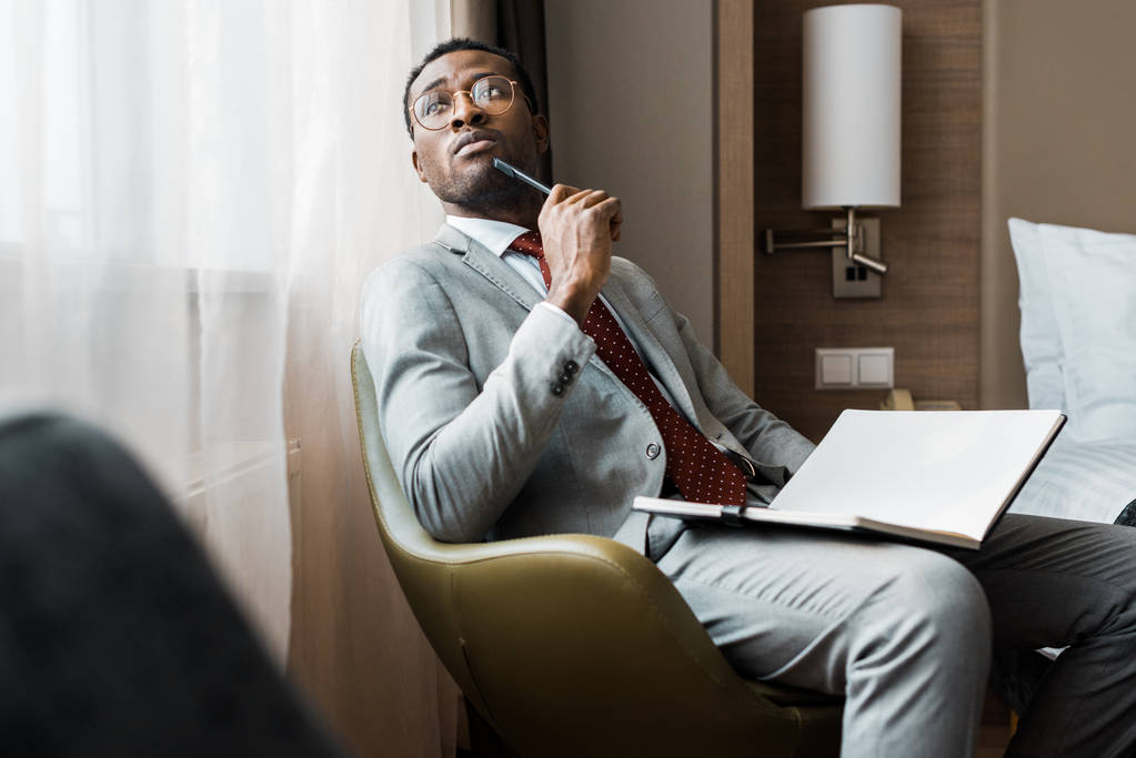 doordachte Afro-Amerikaanse zakenman met dagboek zitten in hotelkamer - Foto, afbeelding