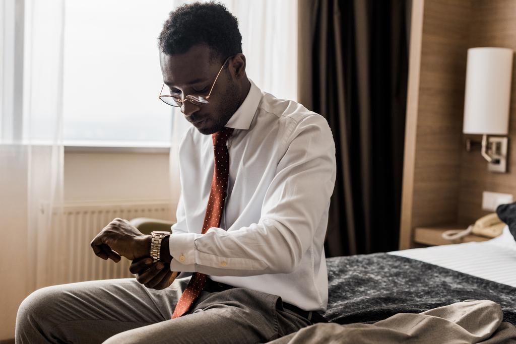 jonge Afro-Amerikaanse zakenman horloge kijken zittend op bed in hotelkamer - Foto, afbeelding
