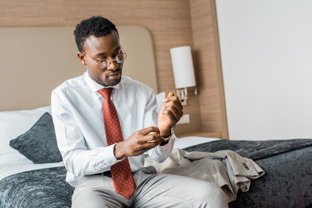 joven hombre de negocios afroamericano con reloj de pulsera sentado en la cama en la habitación de hotel
 - Foto, Imagen