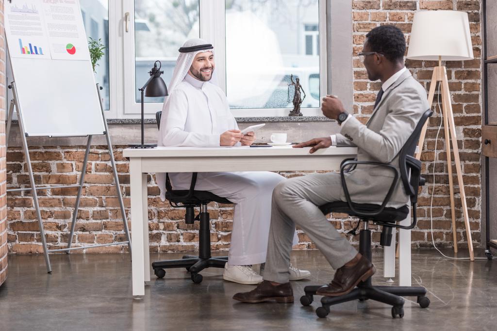 Багатоетнічні бізнесмени розмовляють і посміхаються в сучасному офісі
  - Фото, зображення