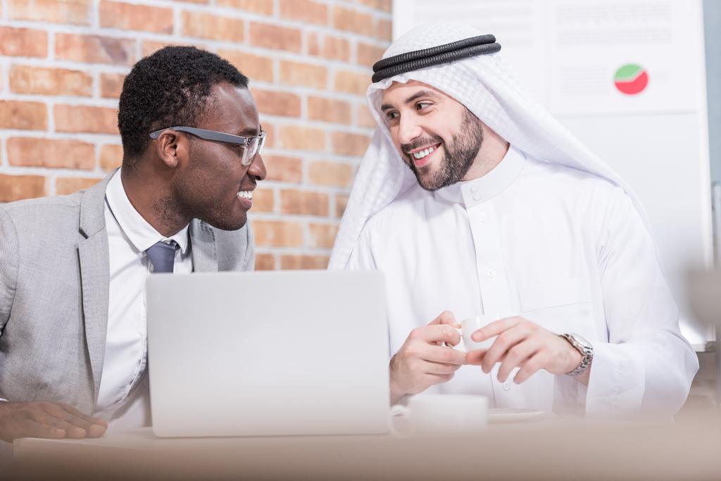 Αραβική επιχειρηματίας κοιτάζοντας αφροαμερικάνων συνεργάτης και χαμογελά στο γραφείο  - Φωτογραφία, εικόνα