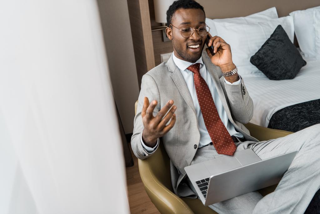onnistunut komea afrikkalainen amerikkalainen liikemies laptop puhuu älypuhelimella hotellihuoneessa
 - Valokuva, kuva
