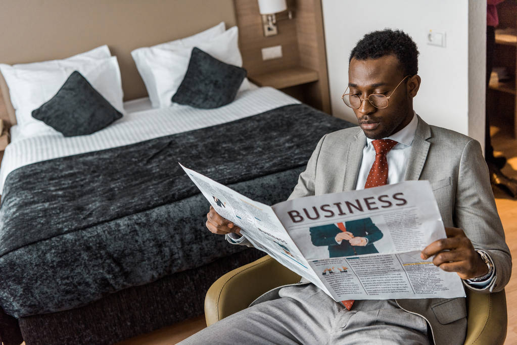 hombre afroamericano serio en traje leyendo periódico de negocios en habitación de hotel
 - Foto, imagen