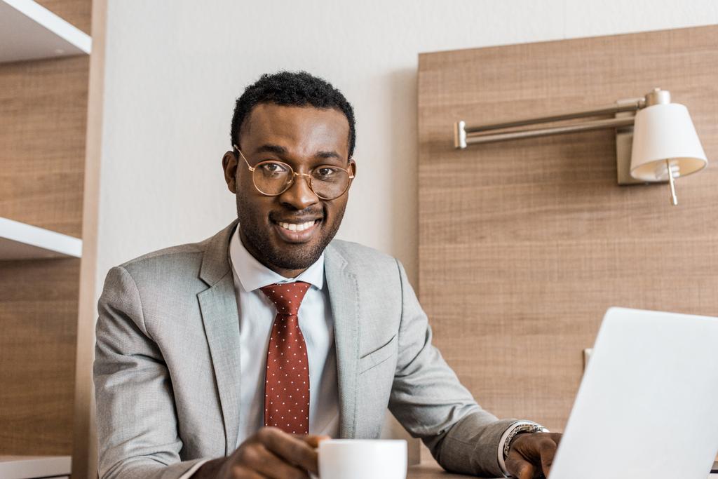 χαμογελώντας Αφρικανικός Αμερικανός επιχειρηματίας με το laptop στο διάλειμμα για καφέ στο δωμάτιο του ξενοδοχείου - Φωτογραφία, εικόνα
