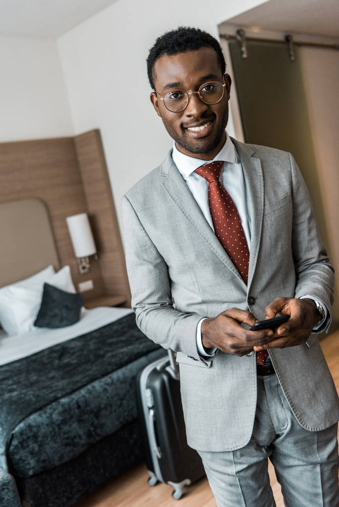 glimlachen van Afro-Amerikaanse zakenman gebruikend smartphone in hotelkamer - Foto, afbeelding