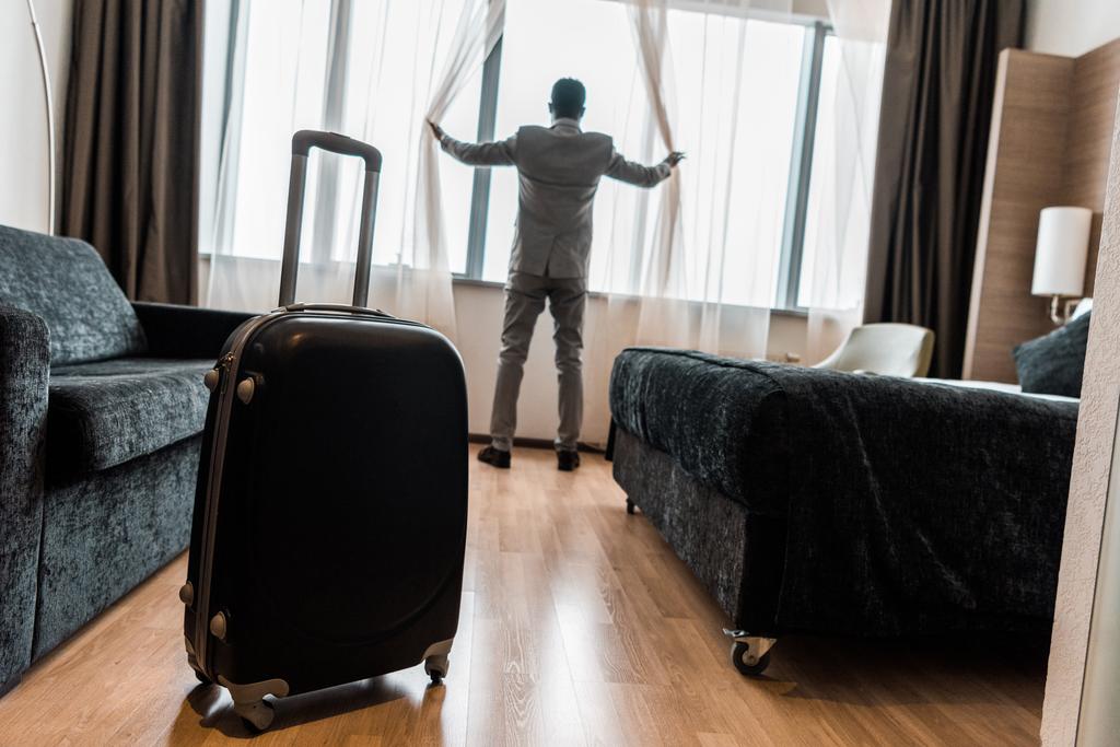 selective focus of suitcase in hotel room with businessman opening window curtains - Photo, Image