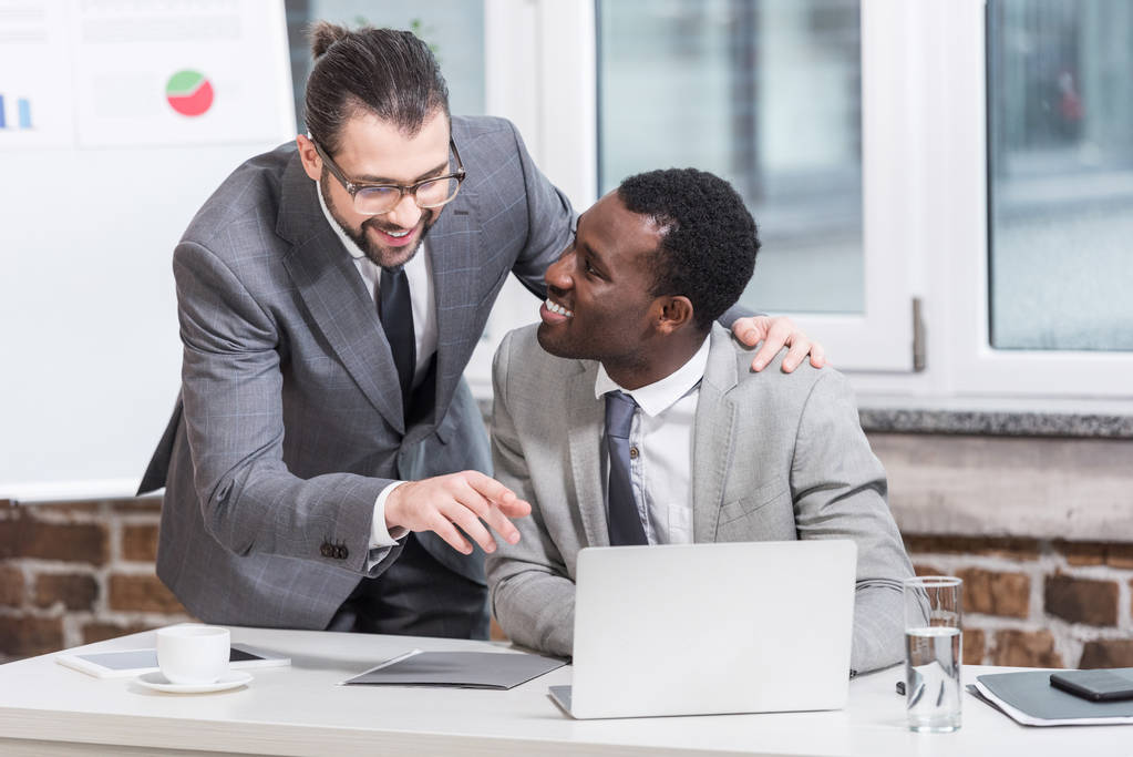 schöner Geschäftsmann umarmt afrikanisch-amerikanischen Partner und zeigt auf Laptop im Büro - Foto, Bild