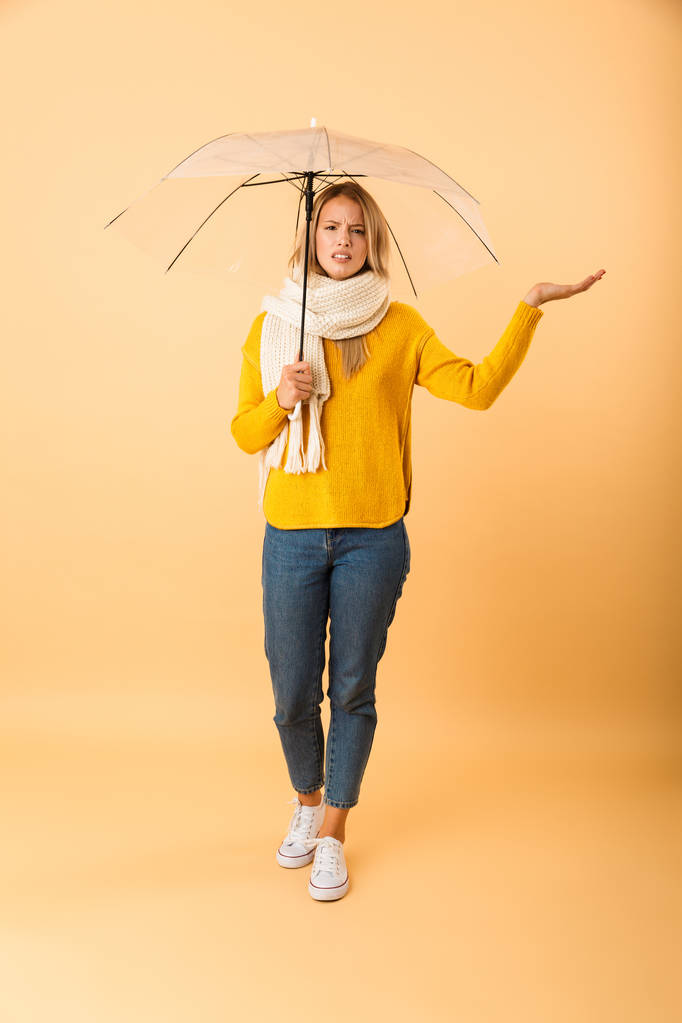 Φωτογραφία: ένα χαριτωμένο γυναίκα που φοράει κασκόλ κρατώντας ομπρέλα θέτοντας απομονωμένη φόντο κίτρινο τοίχο. - Φωτογραφία, εικόνα
