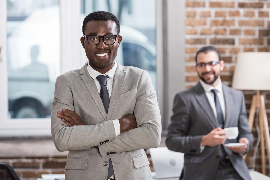 улыбающийся африканский бизнесмен со скрещенными руками, смотрящий в камеру и красивый партнер на заднем плане
 - Фото, изображение