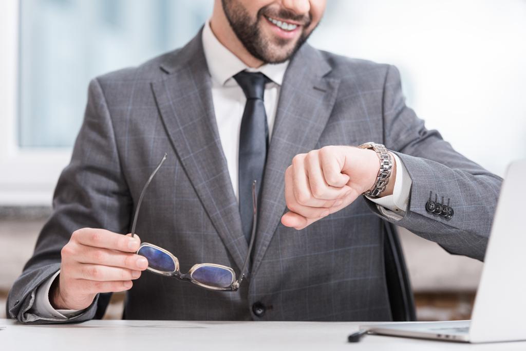 обрезанный вид улыбающегося бизнесмена, держащего очки и смотрящего на часы
 - Фото, изображение