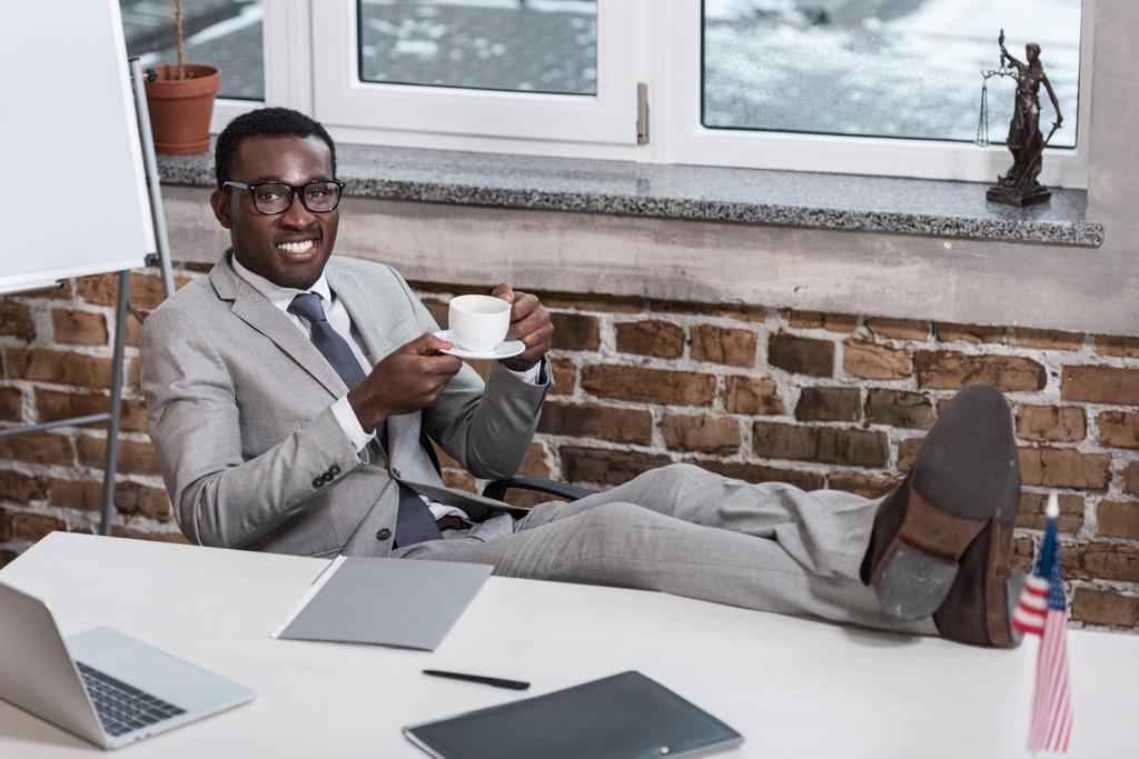 afro-amerikai üzletember asztalnál ül, láb szállt, gazdaság kávéscsésze és látszó-on fényképezőgép irodában mosolyogva - Fotó, kép