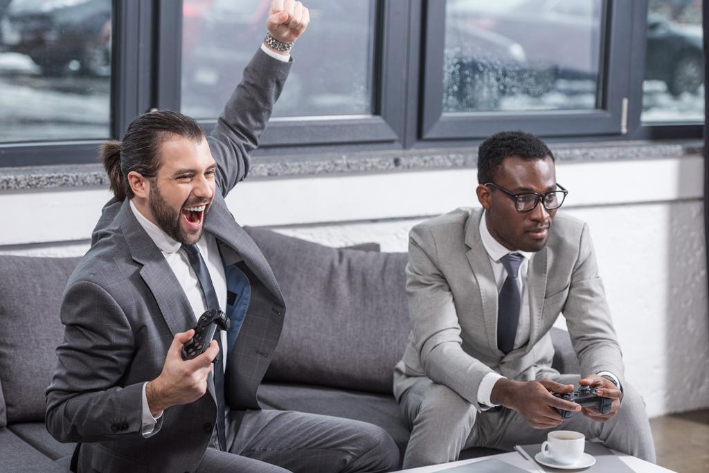 handsome businessman holding gamepad, raising hand and screaming while african american partner looks like loser - Photo, Image