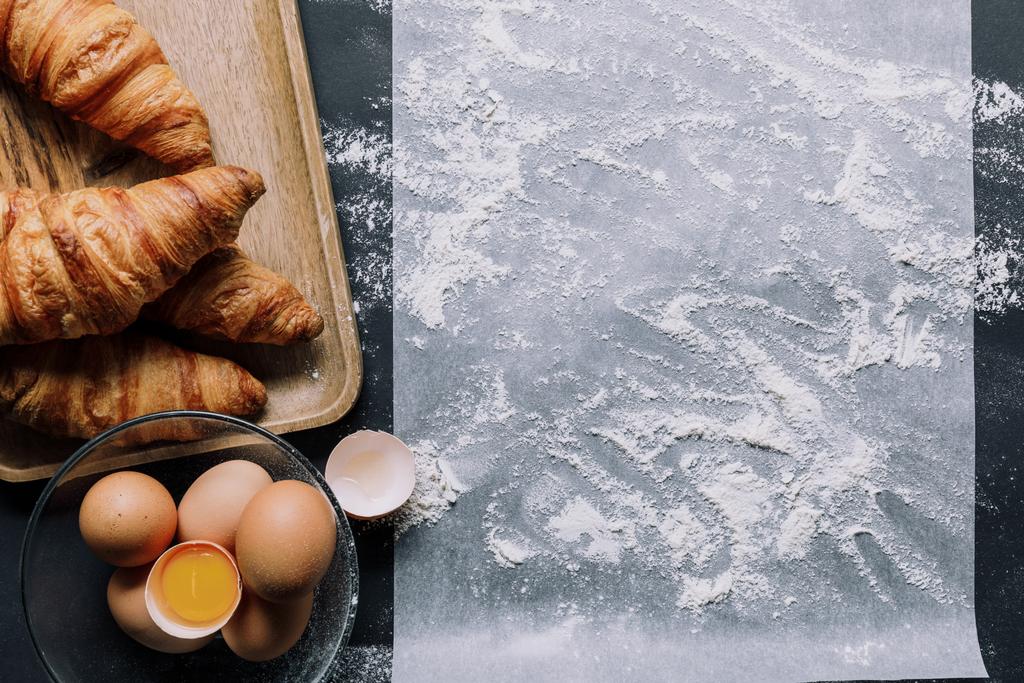 plochou ležela s moukou na papírem na pečení a croissanty s vejci v misce na černý stůl  - Fotografie, Obrázek