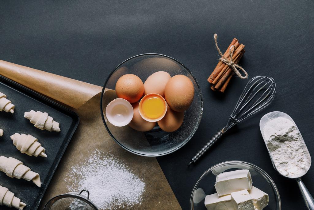 vista superior de los ingredientes, huevos en tazón y bandeja con masa para croissants en mesa negra
 - Foto, imagen
