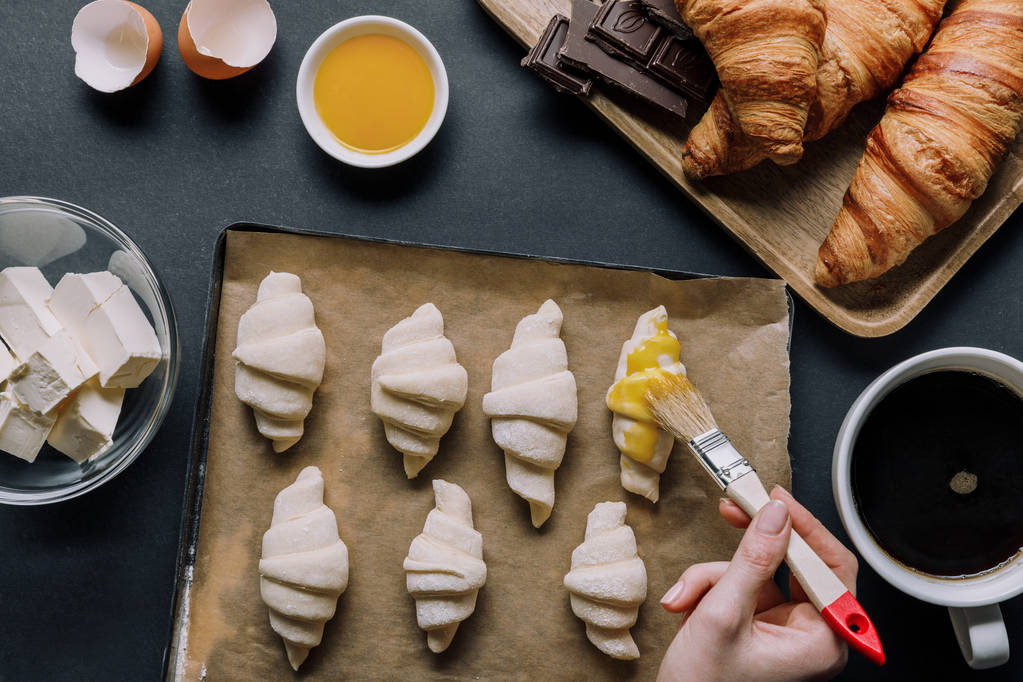 imagem cortada de mulher escovando massa para croissants na bandeja com papel manteiga perto de ingredientes e xícara de café na mesa
 - Foto, Imagem