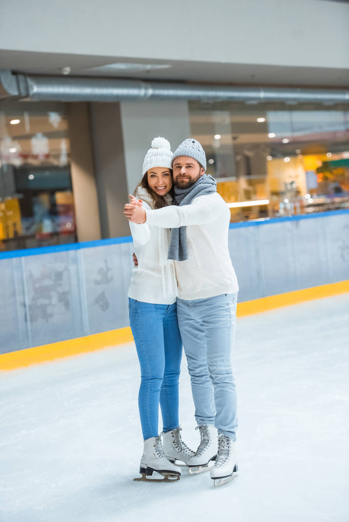 smiling couple in knitted hats and sweaters hugging and holding hands on skating rink - Photo, Image