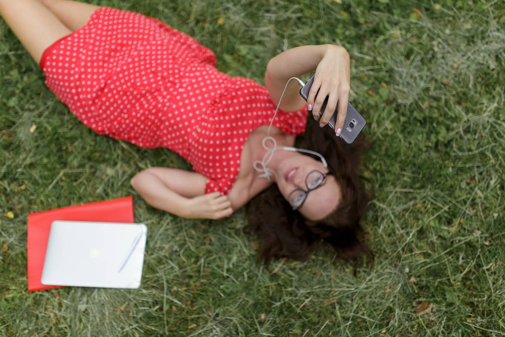 Hermosa mujer toma selfie acostado en la hierba rodeó sus papeles. Vista superior
 - Foto, Imagen