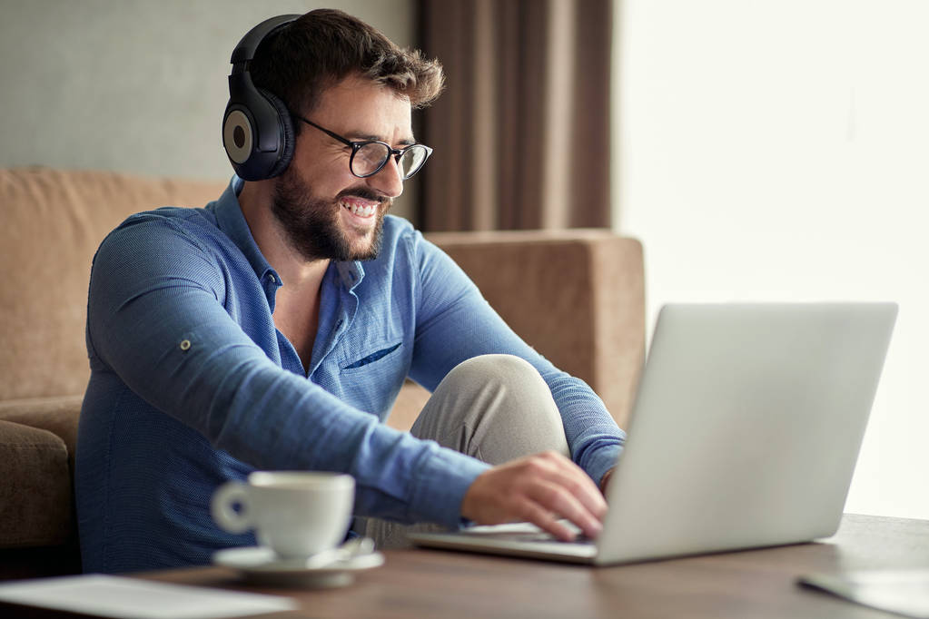 Junger lächelnder Mann mit Kopfhörern mit Laptop in der Wohnung - Foto, Bild