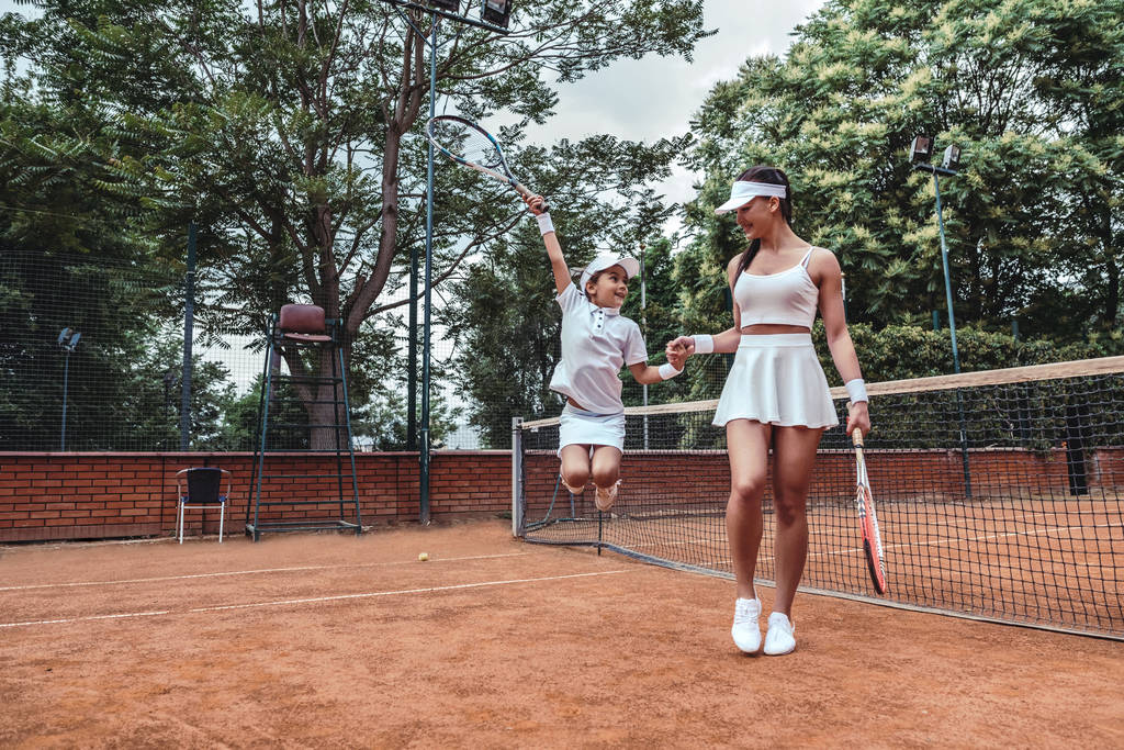 テニスコートで小さなチャンピオン!スポーティなトレーナーと子テニス プレーヤーの跳躍のフルの長さの画像。テニスのコーチは、小さな女の子祝う勝利の手を保持しています。アクティブなレジャー アウトドア. - 写真・画像