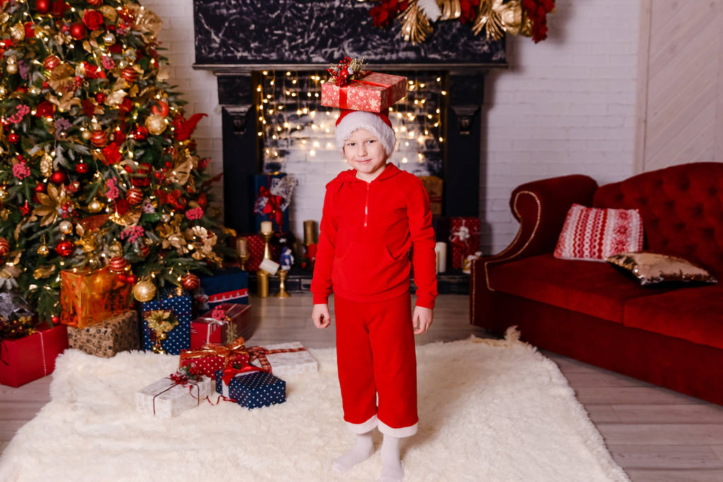 seven-year-old boy in red pants and jacket stands tall with gifts for Christmas, Christmas interior - Photo, Image