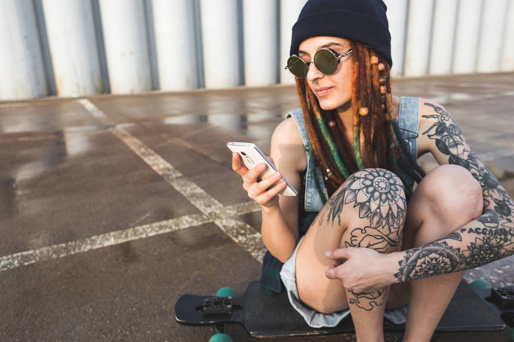 giovane ragazza con tatuaggi e dreadlocks in un cappuccio blu utilizza uno smartphone sullo sfondo di un muro di cemento
 - Foto, immagini