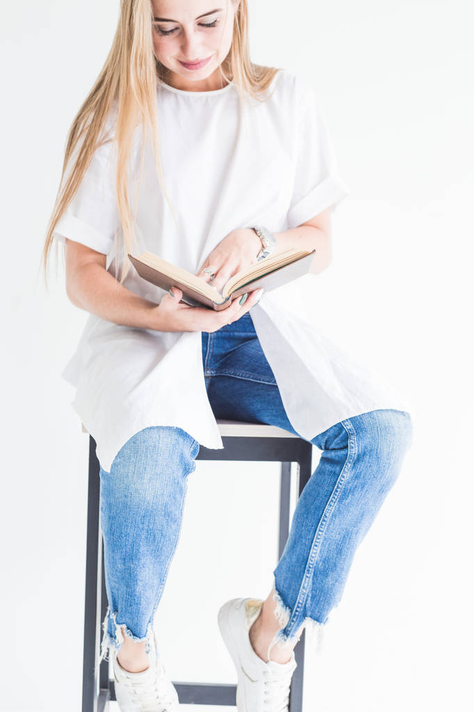 Porträt eines jungen, stylischen blonden Mädchens in weißem T-Shirt und blauer Jeans, das ein Buch auf weißem Hintergrund liest - Foto, Bild