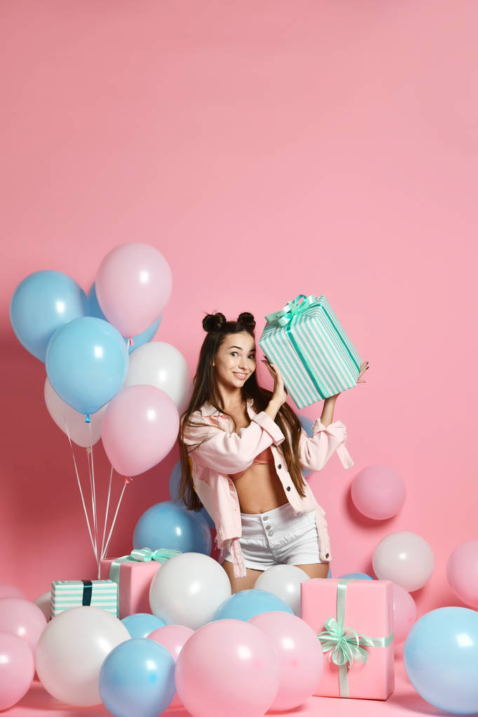 Портрет молодої жінки в день народження, що тримає подарункову коробку з подарунковою коробкою на пастельно-рожевому тлі з різнокольоровими повітряними кульками
.  - Фото, зображення