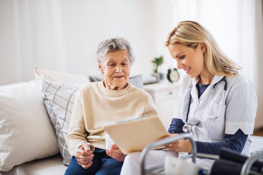 Ošetřovatelkou a starší žena, sedící na posteli v domácím prostředí, mluví. - Fotografie, Obrázek