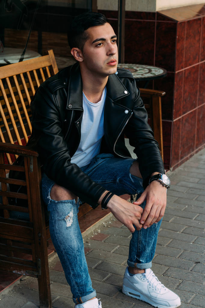aantrekkelijke man zittend op een bankje in een café op de straat, witte sneakers, wit T-shirt, zwart leren jas en jeans, broek. model. wachten voor een vergadering. - Foto, afbeelding