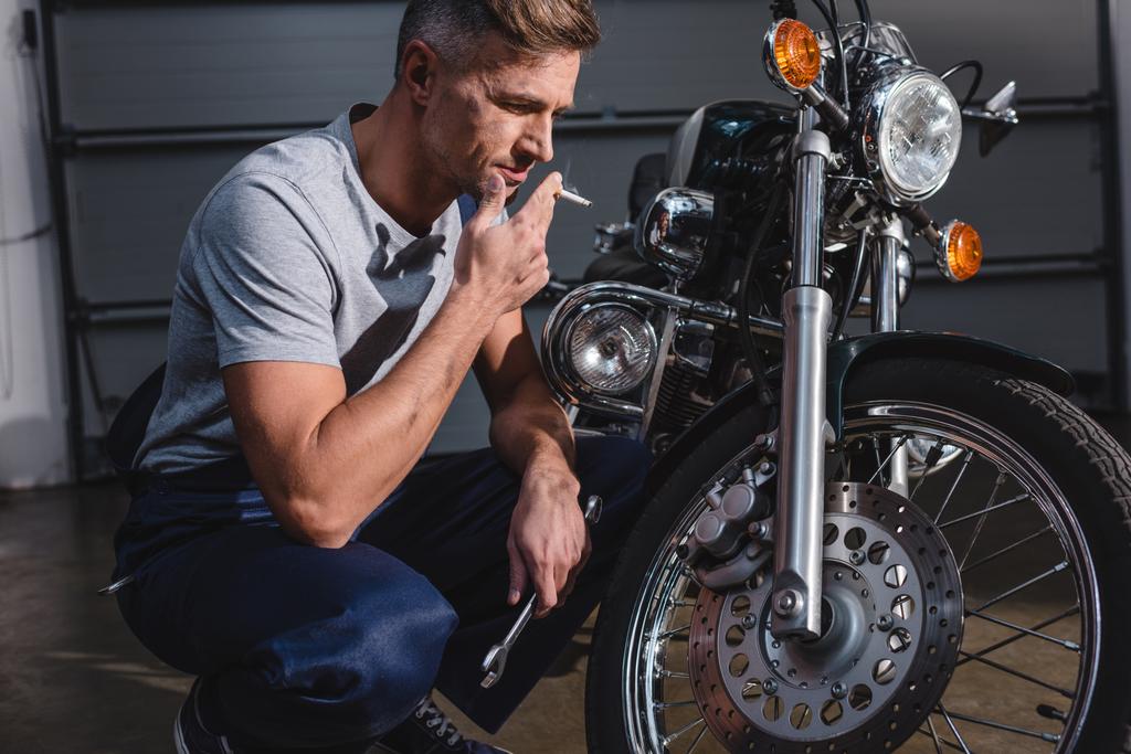 schöner erwachsener Mechaniker raucht und hält Schraubenschlüssel, während er Motorrad in Garage fixiert - Foto, Bild