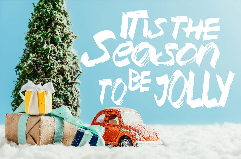 Close-up shot van rode speelgoedauto met presenteert en kerstboom staande op sneeuw gemaakt van katoen op blauwe achtergrond met "het is het seizoen als jolly" inspiratie - Foto, afbeelding