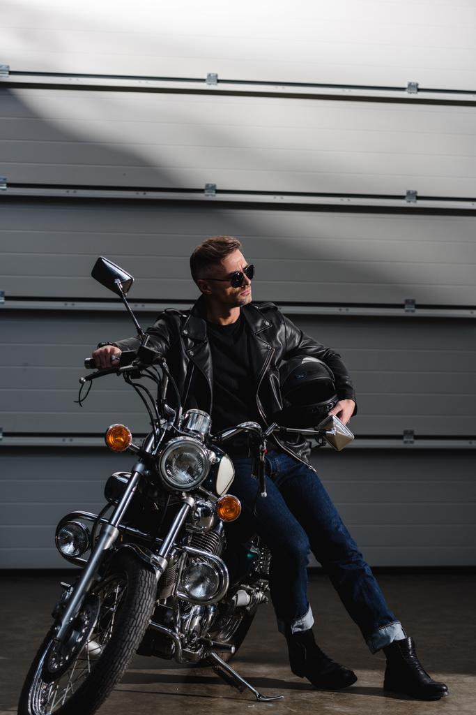 rider clásico en gafas de sol negras y chaqueta de cuero de pie por moto en garaje
 - Foto, imagen