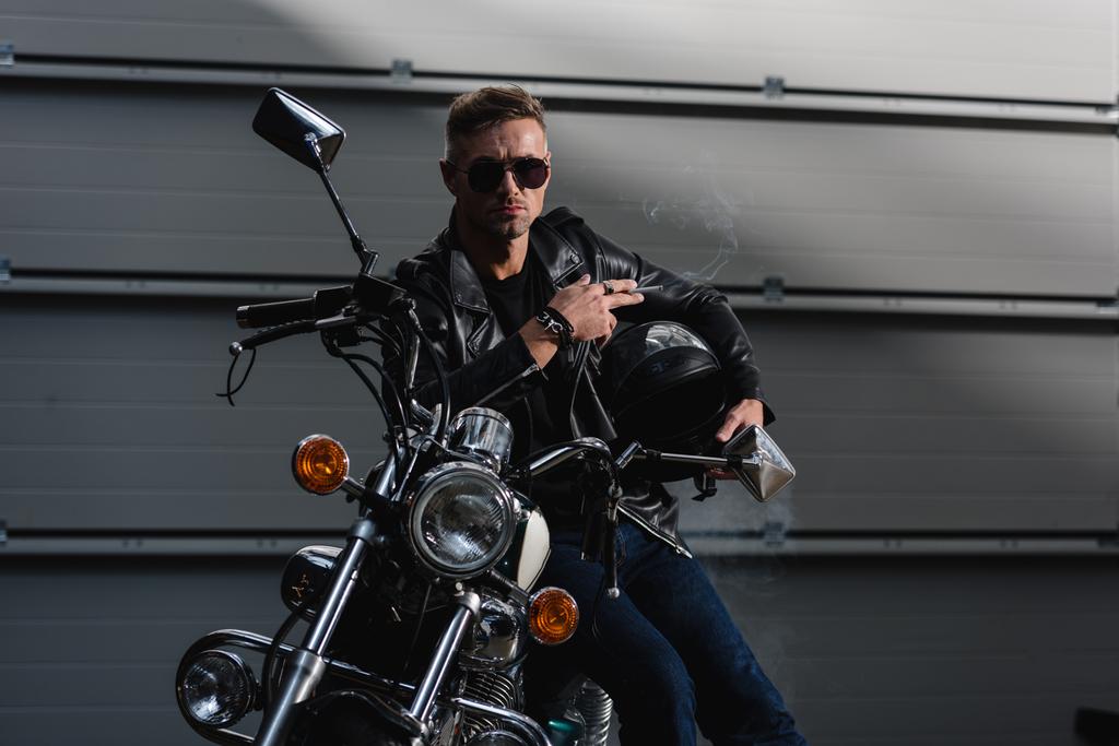 schöner Kerl mit schwarzer Sonnenbrille und Lederjacke sitzt auf Motorrad in Garage - Foto, Bild